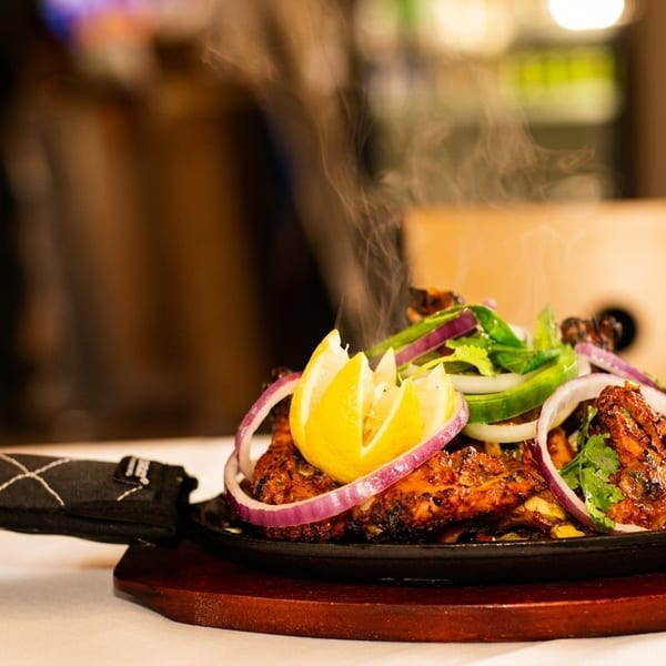 Steaming hot tandoori chicken with lemon, red onion, and green peppers on a sizzling platter.