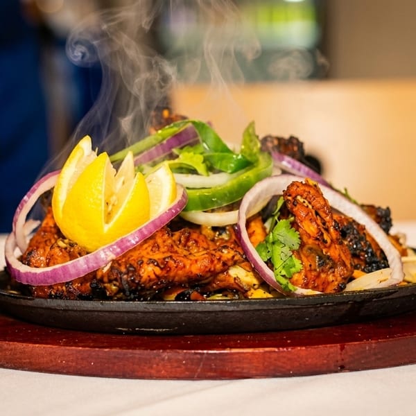 Steaming hot chicken tikka sizzler with onions, bell peppers, and lemon wedges on a wooden platter.