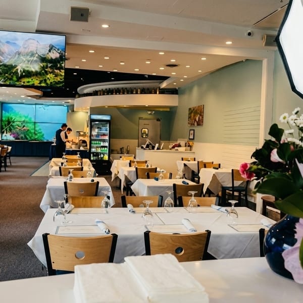 Spacious restaurant dining room with white-clothed tables, blue booths, and a large screen displaying a mountain landscape.
